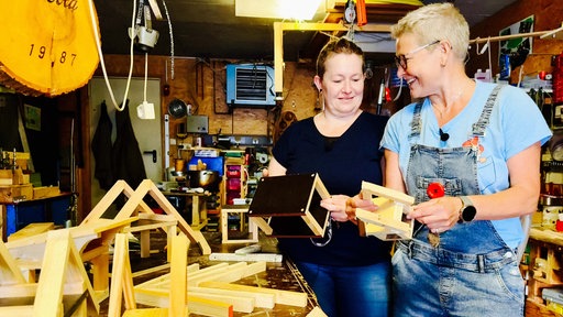 Elke Lohmberg und Annette Lohmberg stehen lachend in einer Wekstatt. Sie halten Holz in der Hand, aus dem ein Wichtelhaus gemacht werden soll.
