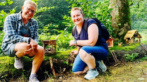 Die Schwestern Elke Lohmberg und Annette Lohmberg knien in einem Wald nebeneinander. Zwischen ihnen steht ein aus Holz gebasteltes kleines Häuschen.