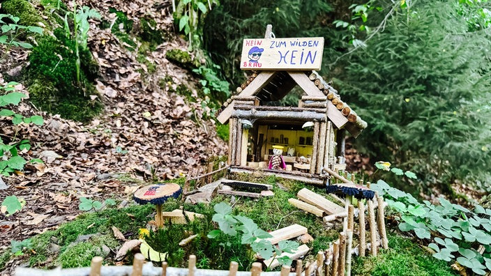 Ein aus Holz gebasteltes Miniatur-Häuschen steht im Wald und stellt einen Wichtel-Biergarten dar.