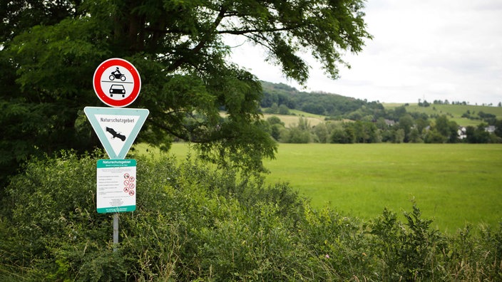 Wiese mit Büschen und Bäumen am Rand, links ein Schild als umgedrehtes Dreieck mit grünem Rand und weißem Grund, darauf steht "Naturschutzgebiet", dazu ein rundes Schild mit rotem Rand und weißenm Grund auf dem ein Motorrad und ein Auto zu sehen sind