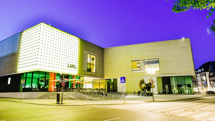Eine abendliche Aufnahme von dem LWL-Museum in Münster: Ein modernes rechteckiges weißes Gebäude mit Lichtinstallation an der linken Wand.