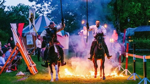 Zwei mittelalterlich gekleidete Männer reiten mit Fackeln in der Hand auf ihren Pferden. Hinter ihnen auf dem Boden brennt ein Feuer. Ein Publikum beobachtet die Show.