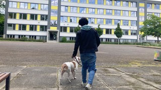 Das Beitragsbild des Dok5 "Ein gutes Leben haben – Jugendliche ohne Schulabschluss" zeigt einen Jugendlichen von hinten mit seinem Hund vor einem Schulgebäude.