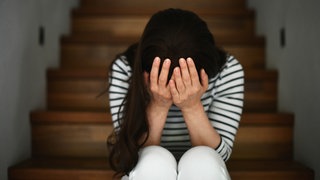 Das Beitragsbild des Dok5 "Erschütterungen – über eine Mutter, die ihr Kind tötet" zeigt eine verzweifelte Frau auf der Treppe sitzend den Kopf in die Hände gelegt. 