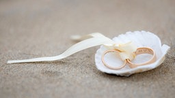 Das Beitragsbild zeigt zwei goldene Eheringe, verbunden mit einem weißen Seidenband in einer weißen Muschel am Sandstrand
