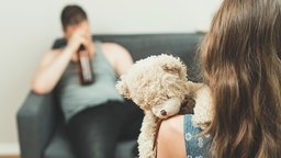 Mädchen mit Plüschteddy steht vor einem Sofa, wo ein betrunkener Mann mit Bierflasche sitzt