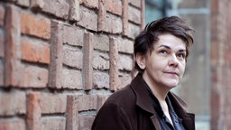 Die Sängerin Christiane Rösinger steht vor einer Backsteinmauer.