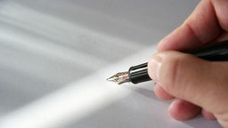 Eine Hand hält einen schwarzen Füller mit Goldfeder und schreibt auf weißem Untergrund, auf den Lichtreflexe fallen.