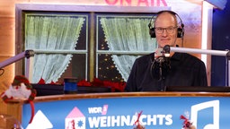 WDR 2 Moderator Thomas Bug trägt Kopfhörer und steht inmitten der WDR 2 Weihnachtswunder-Szenerie, über ihm der "on Air"-Schriftzug