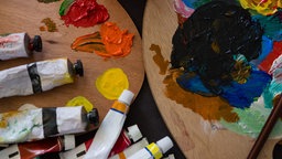 Zwei Farbpaletten mit Farbklecksen, daneben mehrere Tuben Acrylfarbe in verschiedenen Farben