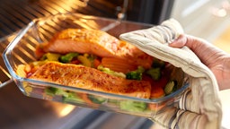 Eine Hand holt gerade mit einem Topflappen eine Auflaufform aus Glas aus dem offen stehenden Ofen. In der Auflaufform befindet sich Gemüse und zwei Stücke Lachs. 