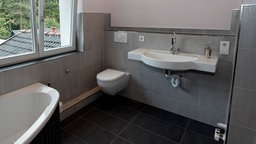 Ein Badezimmer mit grauem Schieferboden. Links ist eine Badewanne, darüber ein Fenster. Mittig ist eine weiße Toilette und ein weißes Spülbecken.