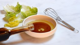 Eine Schale mit Speiseöl und Aceto Balsamico mit einer Flasche Aceto Balsamico, daneben ein Schneebesen und Salatblätter