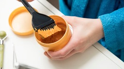 Ein Topf Haarfarbe, von oben fotografiert. Die Person, die die Farbe hält, hält in der anderen Hand einen Haarfärbepinsel, den sie gerade in die Farbe eingetaucht hat. 