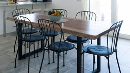 Ein großer Tisch mit Tischplatte aus Holz in einem hellen, modernen Esszimmer. Um den Tisch herum stehen Stühle, auf dem Tisch befindet sich eine Obstschale.