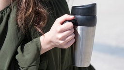 Nahhaufnahme einer Frau, die einen wiederverwendbaren Kaffeebecher mit Griff hält. Der Fokus des Bildes liegt auf dem Becher.