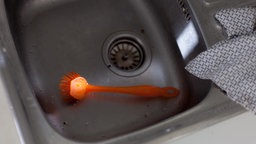 In eine Küchenspüle scheint ein gezielter Sonnenstrahl und beleuchtet eine orangefarbene Spülbürste.