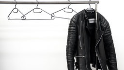 Eine schwarze Lederjacke hängt auf einem Kleiderbügel an einer Kleiderstange. Daran hängen außerdem weitere leere Kleiderbügel. 
