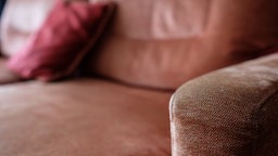 Im Fokus ist die Armlehne eines braunen Sofas, im Hintergrund sieht man das restliche Sofa mit einem roten Kissen