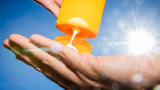 Aus einer orangenen Sonnenmilchtube wird Sonnenmilch auf die Hand gegeben. Im Hintergrund scheint die Sonne an einem strahlend blauen Himmel.