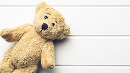 Ein kleiner, heller Teddybär auf einem weißen Holzuntergrund. 