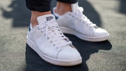 Weiße Sneaker mit weißen Schnürsenkeln. Es ist der Ansatz der Knöchel und einer grauen Hose zu sehen.