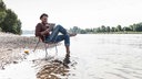 Symbolbild: Ein Mensch sitzt lächelnd mit einem Tablet alleine auf einem Campingstuhl am Wasser.