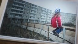 Ein kleines Mädchen sitzt auf einem Holzgeländer eines Spielplatzes im Innenhof einer Plattenbau Siedlung.