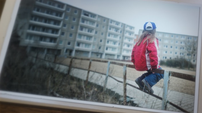 Ein kleines Mädchen sitzt auf einem Holzgeländer eines Spielplatzes im Innenhof einer Plattenbau Siedlung.