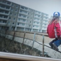 Ein kleines Mädchen sitzt auf einem Holzgeländer eines Spielplatzes im Innenhof einer Plattenbau Siedlung.