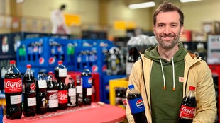 Das Bild zeigt Daniel Aßmann mit verschiedenen Colamarken auf einem Tisch. In der Hand hält er jeweils zwei Wasserflaschen. 