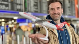 Daniel Aßmann ist am Flughafen und hält ein Modellflugzeug lächelnd in die Kamera.
