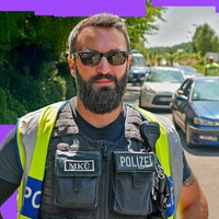 Ein Polizist in Uniform und Sonnenbrille blickt in die Kamera. Hinter ihm eine Schlange an Autos