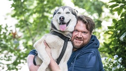 Das Bild zeigt einen Hund angekuschelt an einen sitzenden Mann. 