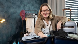 Das Bild zeigt Yvonne Willicks, vor ihr ein offener Koffern. Im Hintergrund ist eine Kleiderstange zu sehen.