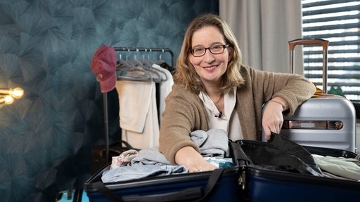 Das Bild zeigt Yvonne Willicks, vor ihr ein offener Koffern. Im Hintergrund ist eine Kleiderstange zu sehen.