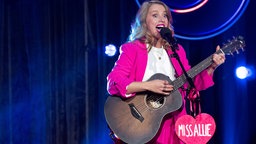 Miss Allie steht in einem pinken Hosenanzug und mit einer Gitarre auf der Bühne