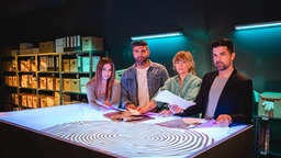 Das Mordorte-Team steht um einen Tisch herum und blickt in die Kamera