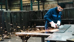 Ein Azubi mit Schutzbrille schleift eine Metallplatte, dabei fliegen viele Funken