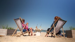 Zwei Personen sitzen in Liegestühlen am Strand. 