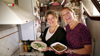 Tamina und eine andere Frau stehen in der Küche und präsentieren ihr Gericht.