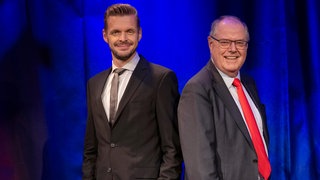 Zwei Männer in Anzug und Krawatte stehen Schulter an Schulter vor einem dunkelblauen Vorhang und blicken freundlich in die Kamera