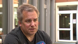 Man sieht einen Mann in schwarzen T-Shirt vor einem gläsernden Gebäude. 