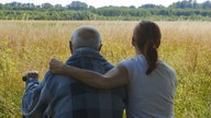 Ein älterer Herr sitzt mit seiner Tochter auf einer Bank vor einem großen Feld.
