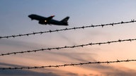 Flugzeug vor Abendhimmel, davor Stacheldrahtzaun