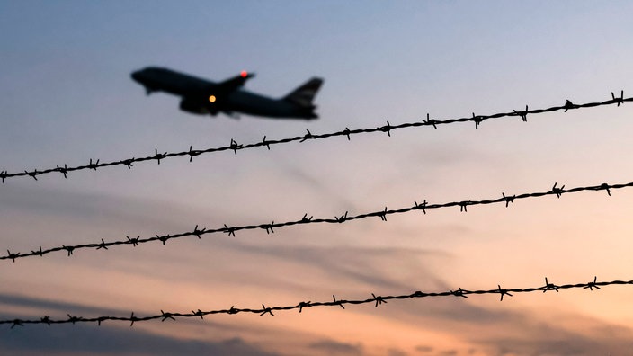 Flugzeug vor Abendhimmel, davor Stacheldrahtzaun