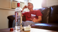 Ein Mann sitzt auf dem Sofa, vor sich eine Flasche Schnaps und Zigaretten