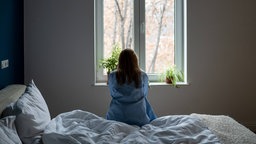 Eine Frau sitzt alleine auf einem Bett und schaut aus dem Fenster