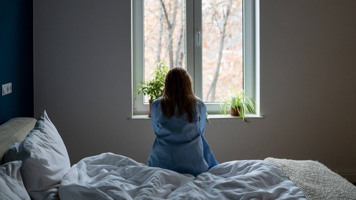 Eine Frau sitzt alleine auf einem Bett und schaut aus dem Fenster