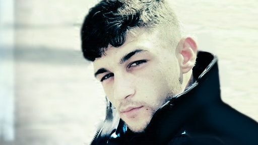 Ein junger Mann in der Nahaufnahme. Er trägt einen modischen Kurzhaarschnitt und hat den Kragen seines Anoraks lässig aufgestellt.